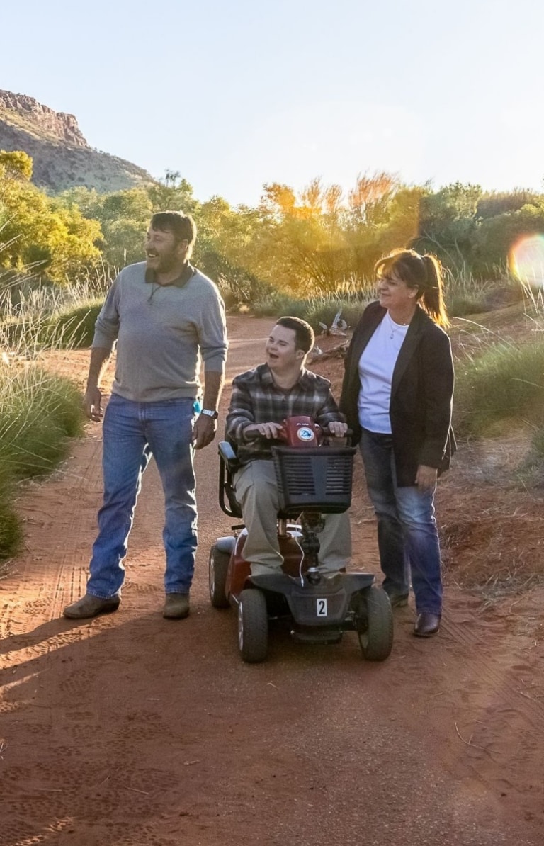 Man seated on a mobility device with another man and woman, wandering among the red dirt and greenery of Alice Springs Desert Park with the sun in the background in Alice Springs, Northern Territory © Tourism NT/Helen Orr Man seated on a mobility device with another man and woman, wandering among the red dirt and greenery of Alice Springs Desert Park with the sun in the background in Alice Springs, Northern Territory © Tourism NT/Helen Orr