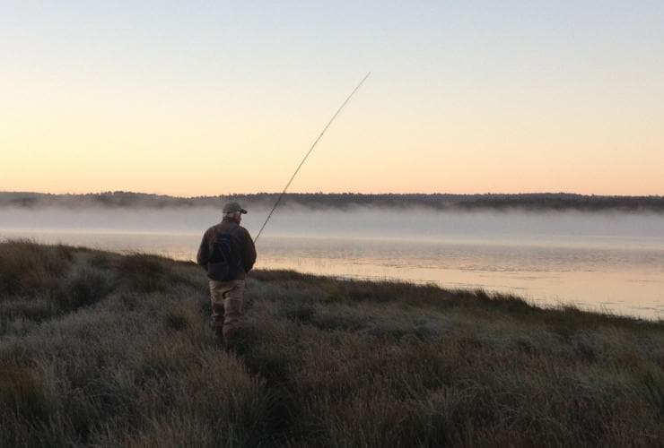 Ein Mann beim Angeln am Ufer eines von Nebelschwaden bedeckten Sees bei Sonnenuntergang, Central Highlands, Tasmanien © Driftwater