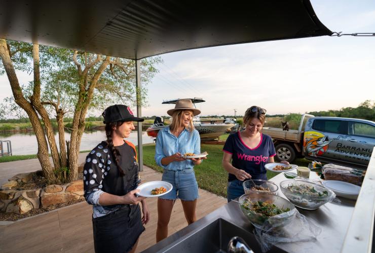 Tre persone che si servono cibo con un corso d'acqua e una barca sullo sfondo con Barramundi Adventures Darwin, Berry Springs, Northern Territory © Tourism Australia